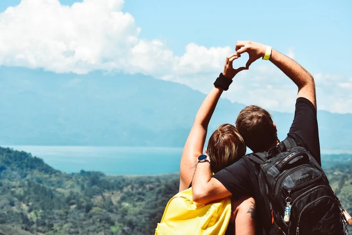 A couple with backpacks forming a heart shape with their hands, symbolizing love and adventure.